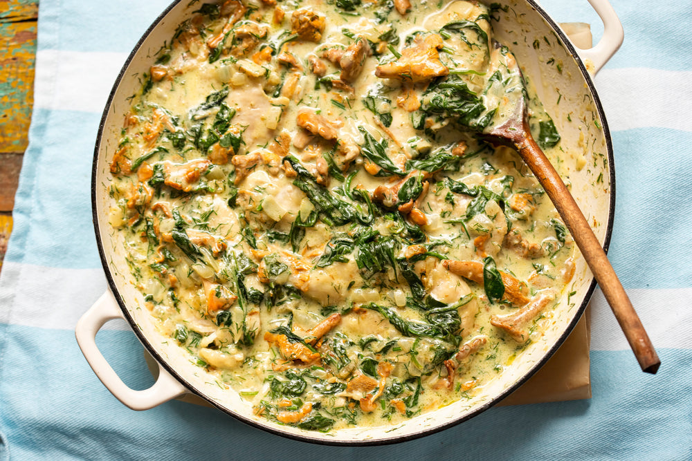 A creamy spinach casserole with sautéed mushrooms and herbs is served in a round dish, with a wooden spoon resting inside, placed on a light blue striped cloth.