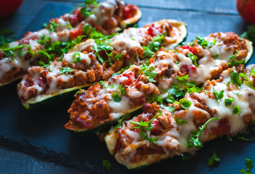 Stuffed zucchini halves are topped with melted cheese and fresh herbs, arranged on a dark slate surface, showcasing a delicious and colorful filling of meat and vegetables.