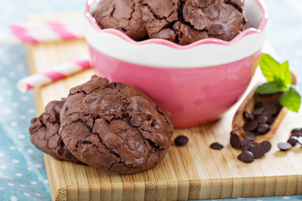 Chocolate meringue cookies are arranged on a wooden board beside a pink bowl filled with more cookies, with chocolate chips scattered around and a sprig of mint for decoration.