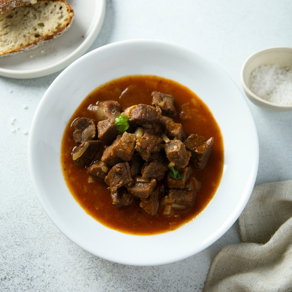 A white bowl filled with tender, brown Swiss steak chunks in a rich tomato sauce is placed on a light surface, accompanied by a slice of bread and a small bowl of salt.