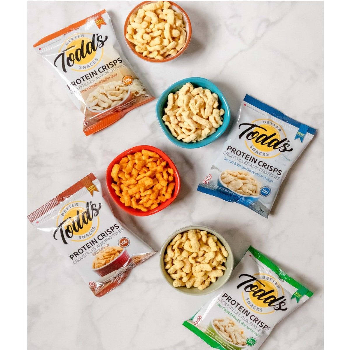 Three bags of Todd's Better Snacks Protein Crisps are displayed alongside colorful bowls filled with different flavors of the snacks, all arranged on a light marble surface.