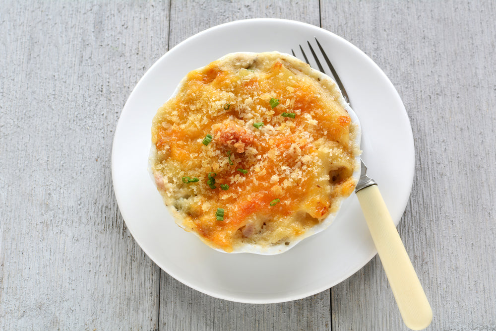 A baked tuna casserole topped with golden breadcrumbs and green herbs sits on a white plate, accompanied by a fork, against a light wooden background.