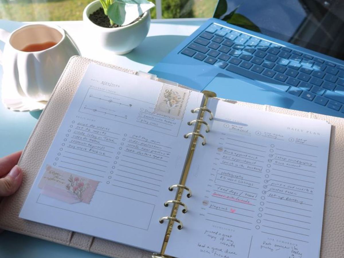 A person holds an open planner filled with notes and to-do lists, accompanied by a cup of tea and a laptop, in a bright, sunlit setting, suggesting preparation for bariatric surgery.