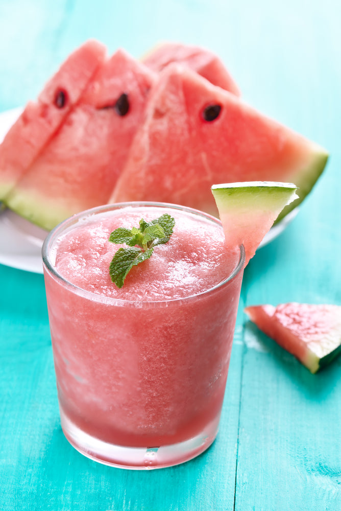 A refreshing watermelon smoothie with a mint garnish is served in a clear glass, accompanied by slices of watermelon on a white plate, all set against a bright turquoise background.