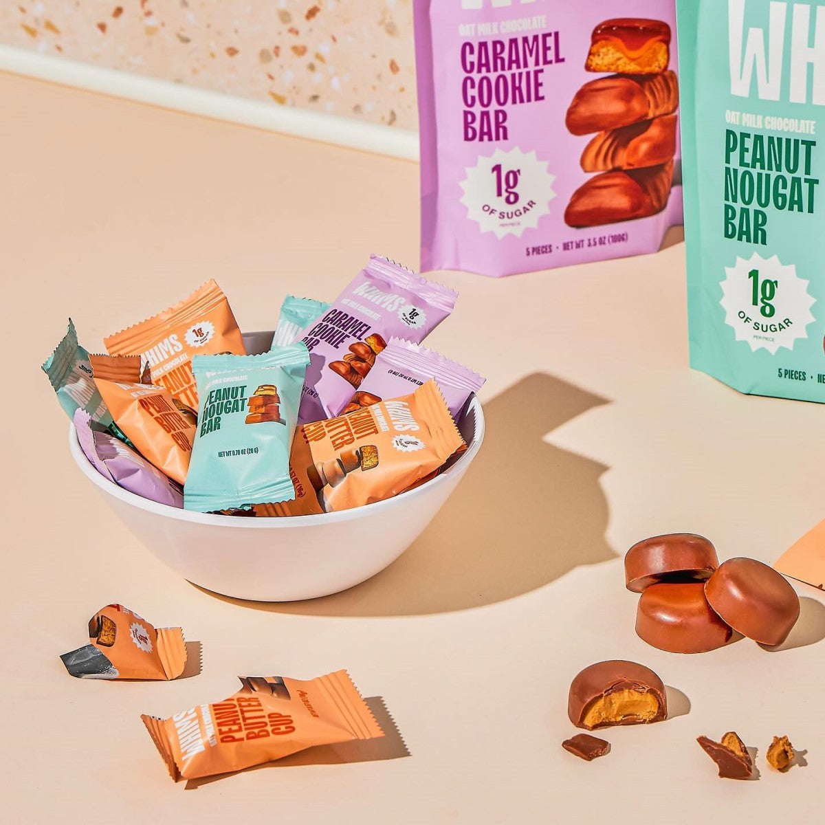 A white bowl filled with colorful snack wrappers of WHIMS Oat Milk Chocolate Bars sits on a light surface, with a few unwrapped bars and packaging of Caramel Cookie and Peanut Nougat bars in the background.