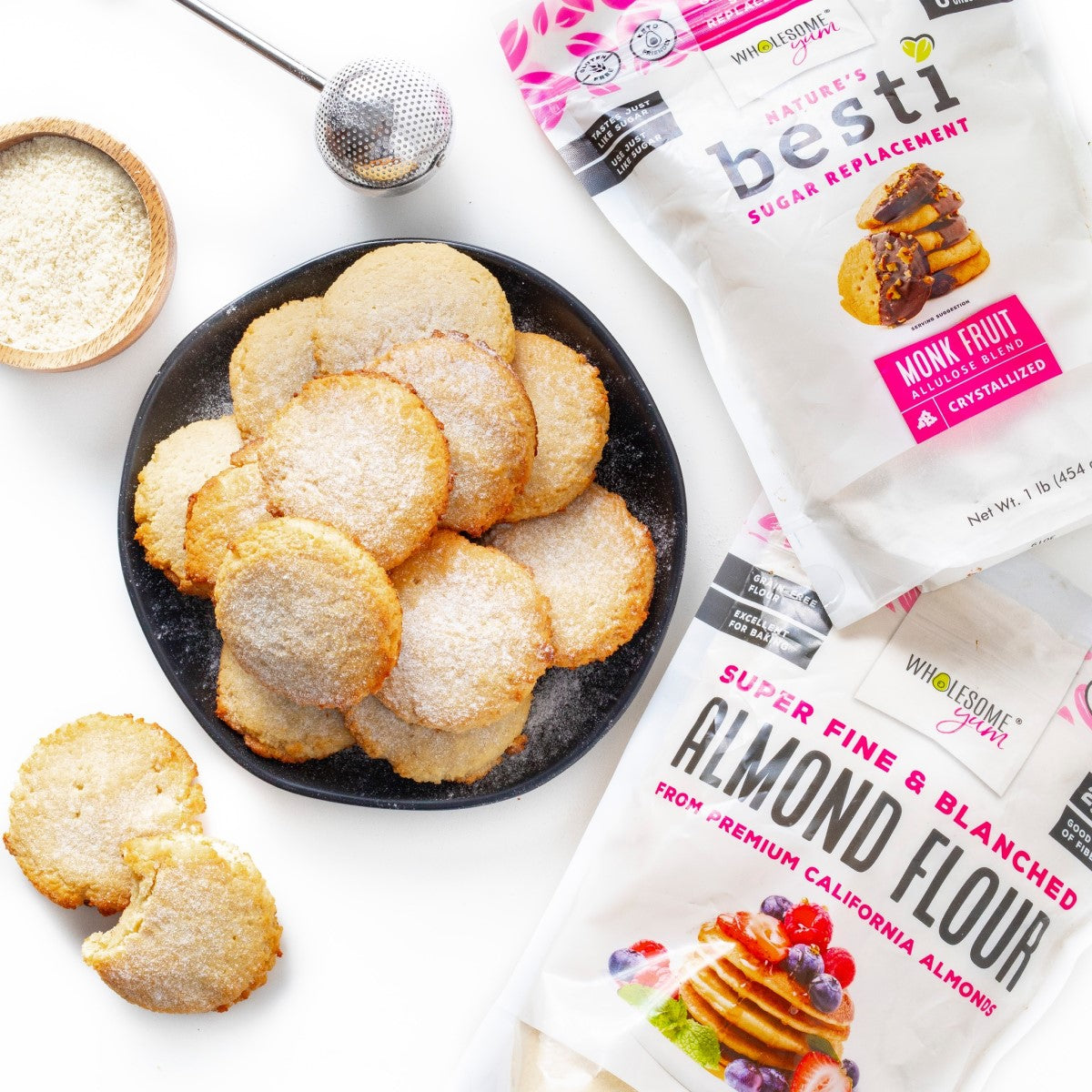 A black plate holds several golden-brown cookies dusted with sugar, alongside bags of almond flour and sugar replacement, with a small bowl of flour and a measuring tool in the background.