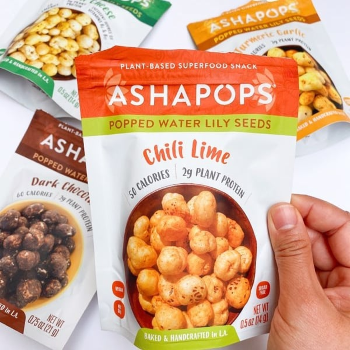 A hand holds a package of "Chili Lime" popped water lily seeds, featuring a colorful design, while other snack varieties are visible in the background, highlighting a healthy snack option.