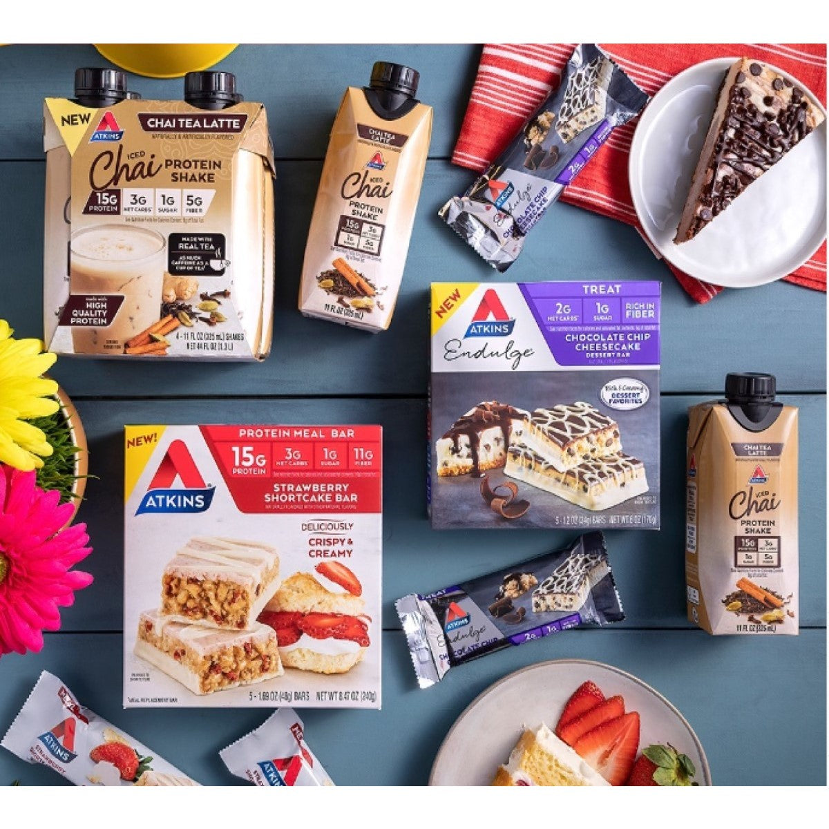 Atkins products are displayed on a blue wooden surface, featuring new chai protein shakes, protein meal bars, and a chocolate chip cheesecake dessert, surrounded by colorful flowers and a slice of cheesecake.