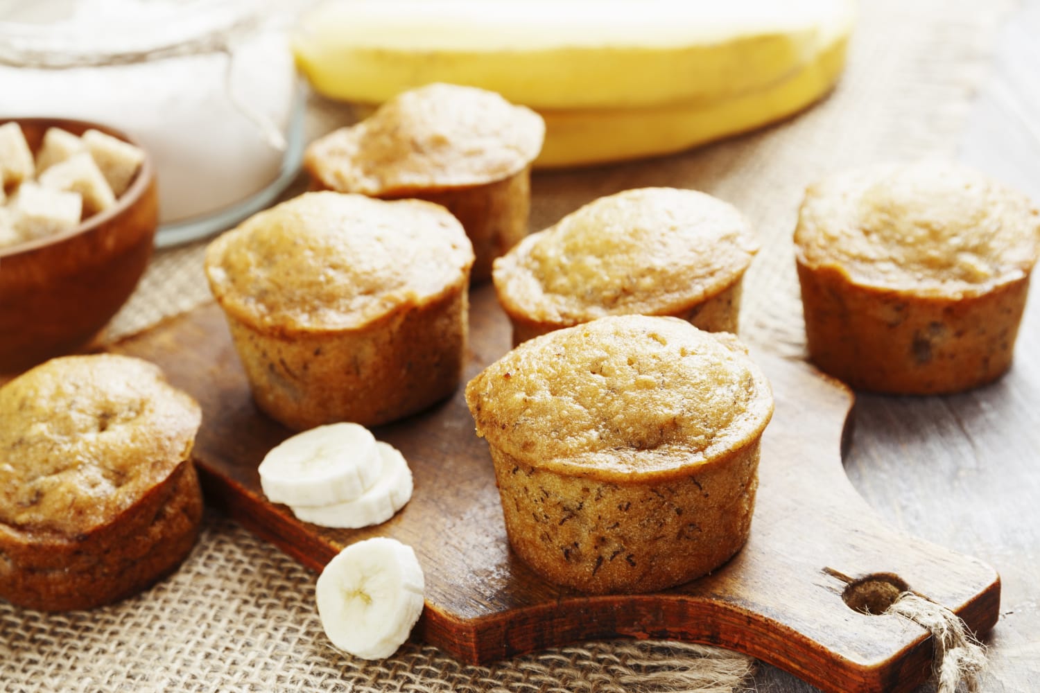 Six golden-brown banana muffins are arranged on a wooden cutting board, with sliced bananas and a bowl of sugar in the background, creating a cozy baking scene.