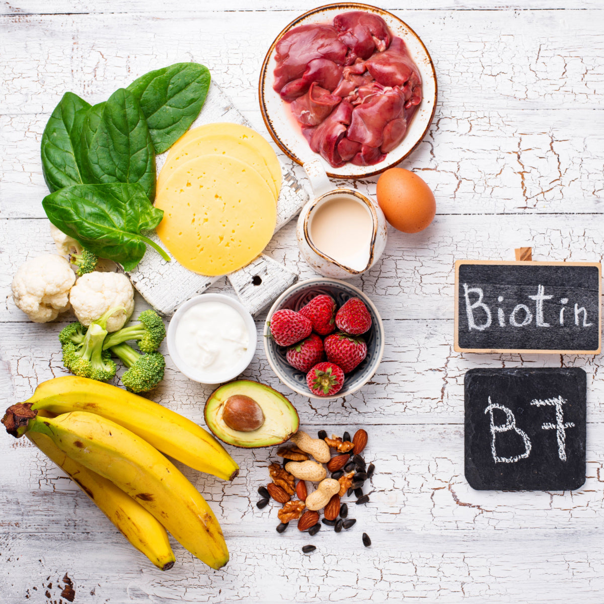 A variety of foods rich in biotin, including spinach, eggs, strawberries, bananas, and nuts, are arranged on a rustic wooden surface, with small chalkboards labeled "Biotin" and "B7."