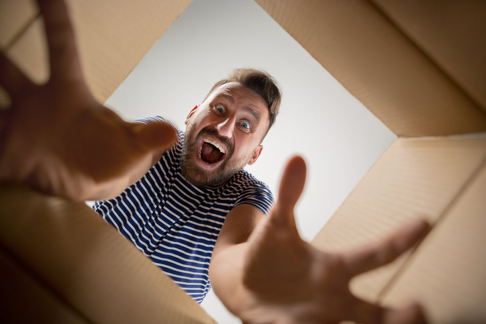 A man with a surprised expression reaches excitedly into a cardboard box, as if unveiling its contents, with a neutral background highlighting his enthusiasm.