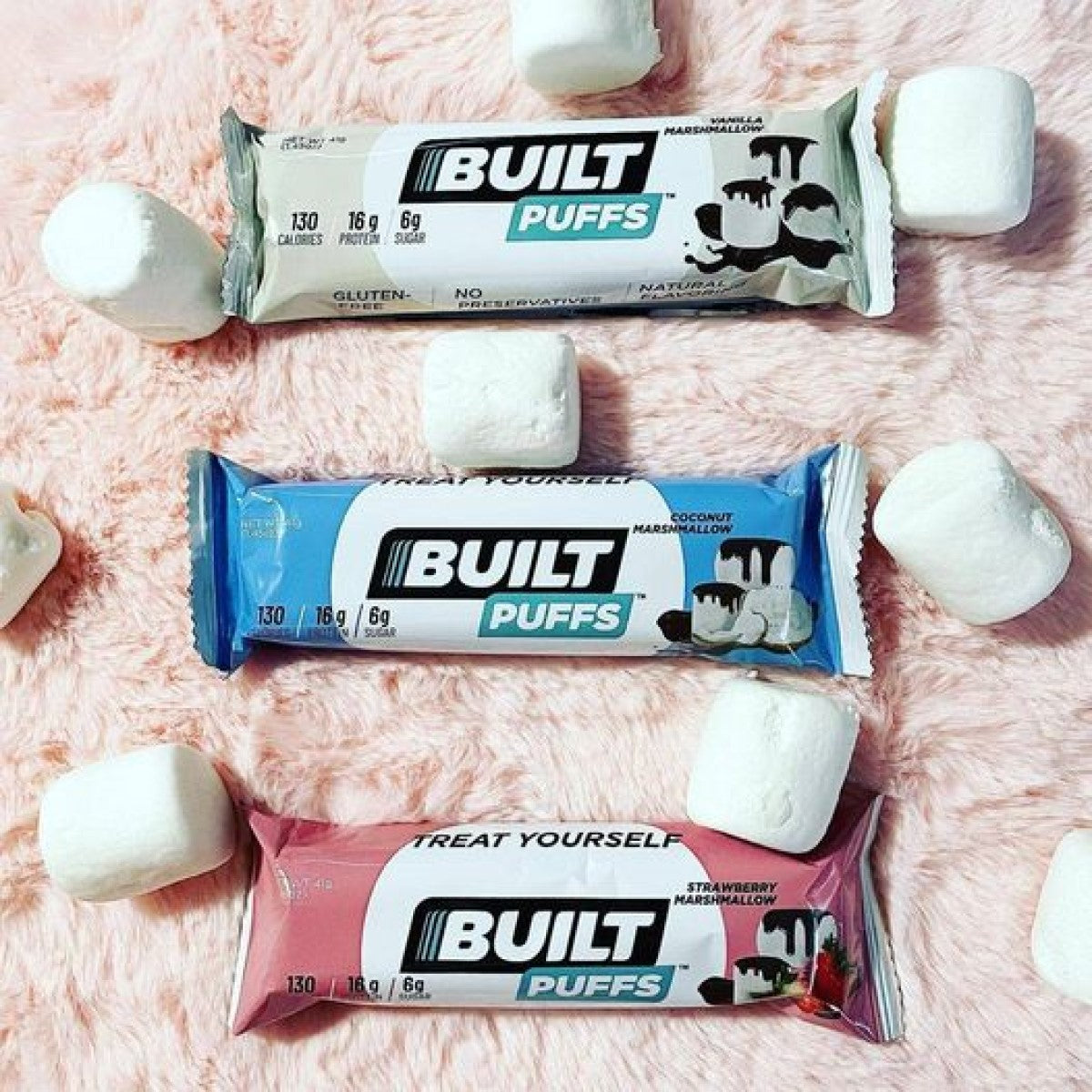Three Built Bar Protein Puffs in vanilla, coconut, and strawberry flavors are displayed on a soft pink surface, surrounded by fluffy marshmallows.