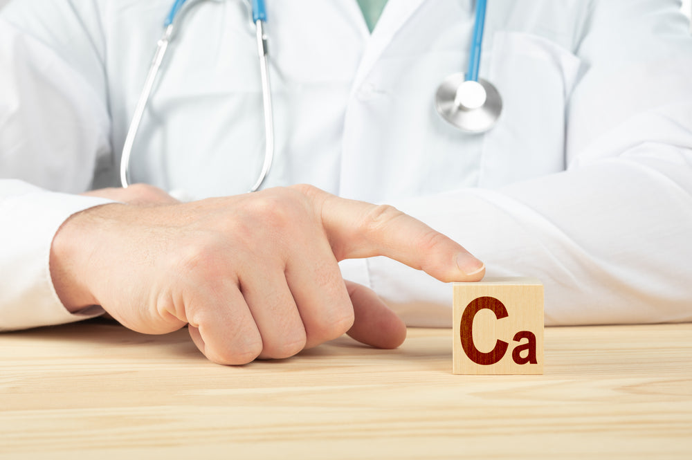 A healthcare professional in a white coat points to a wooden block displaying "Ca," symbolizing calcium, on a wooden table, emphasizing its importance post-bariatric surgery.