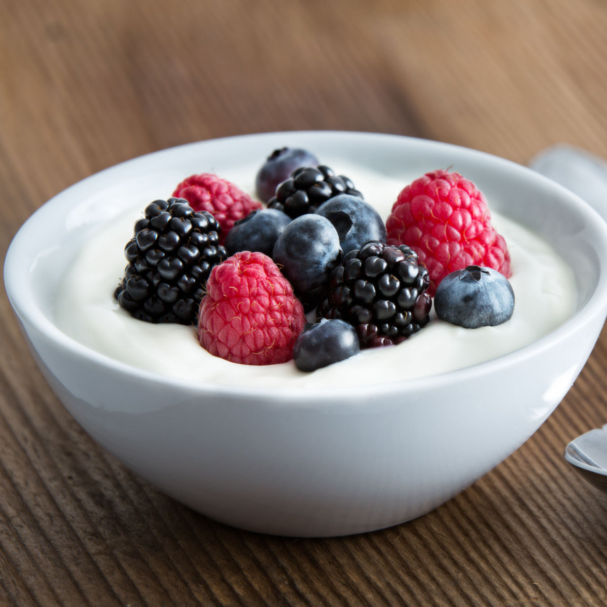 A white bowl filled with creamy yogurt topped with fresh raspberries, blackberries, and blueberries sits on a wooden surface, highlighting a colorful and healthy dessert option.