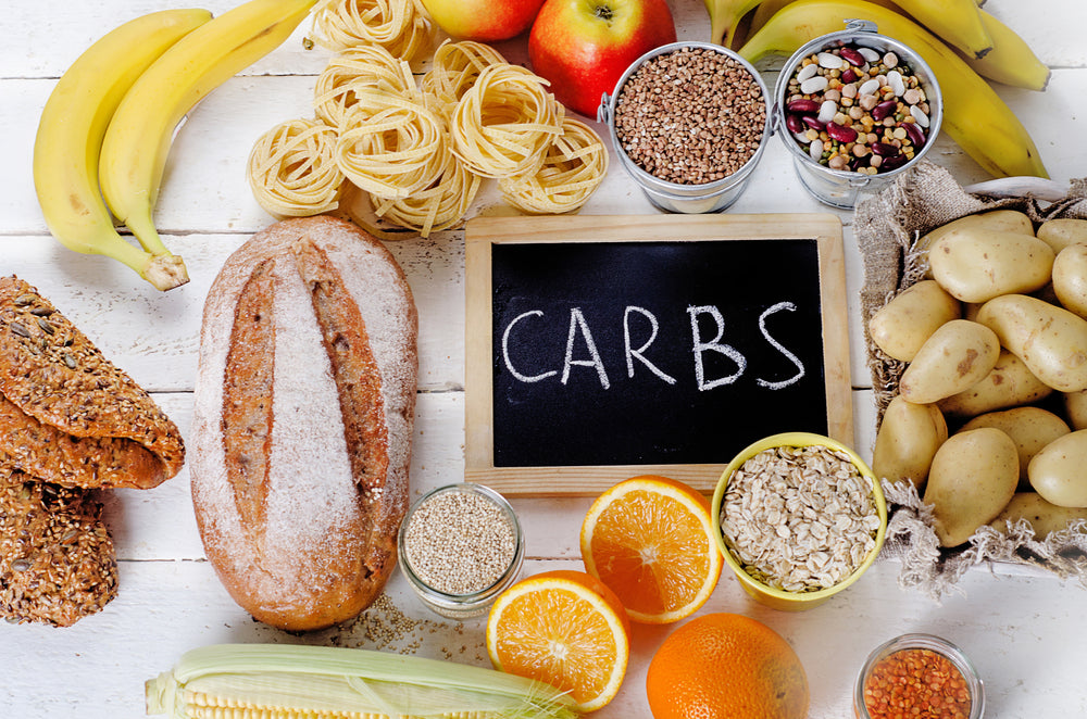 A variety of carbohydrate-rich foods, including bread, pasta, fruits, and grains, are arranged around a chalkboard labeled "CARBS" on a wooden surface.