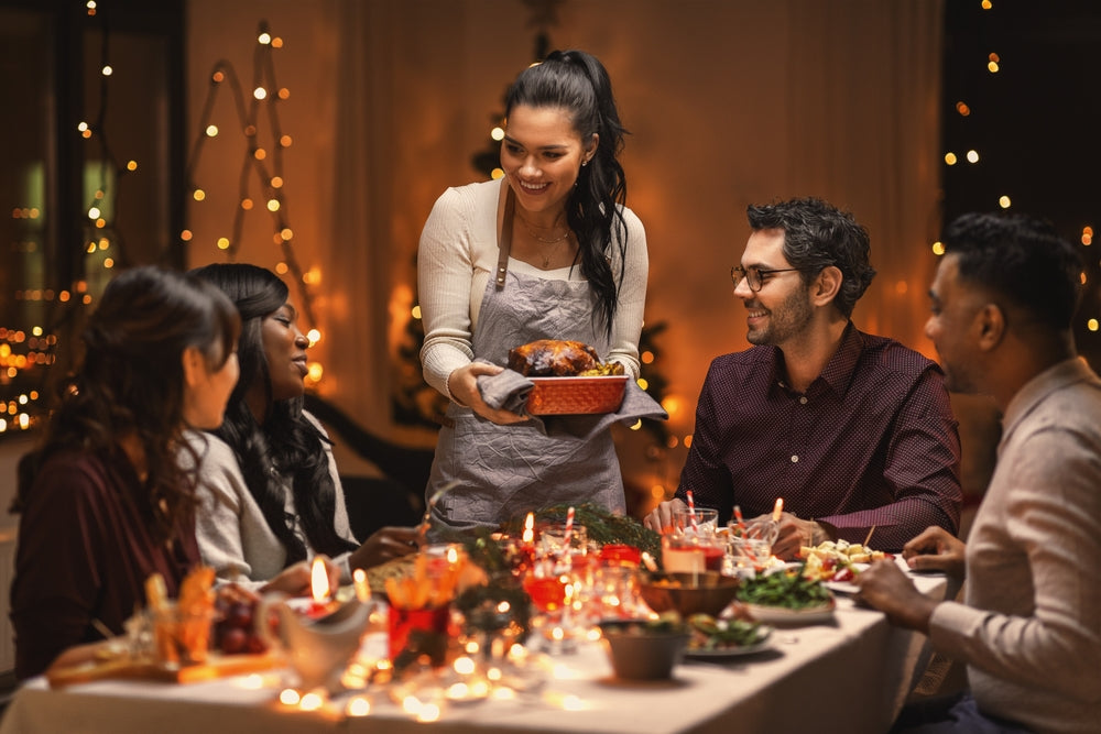 A woman in an apron serves a roasted dish to a group of smiling friends at a festive dinner table adorned with candles and holiday decorations, creating a warm and joyful atmosphere.