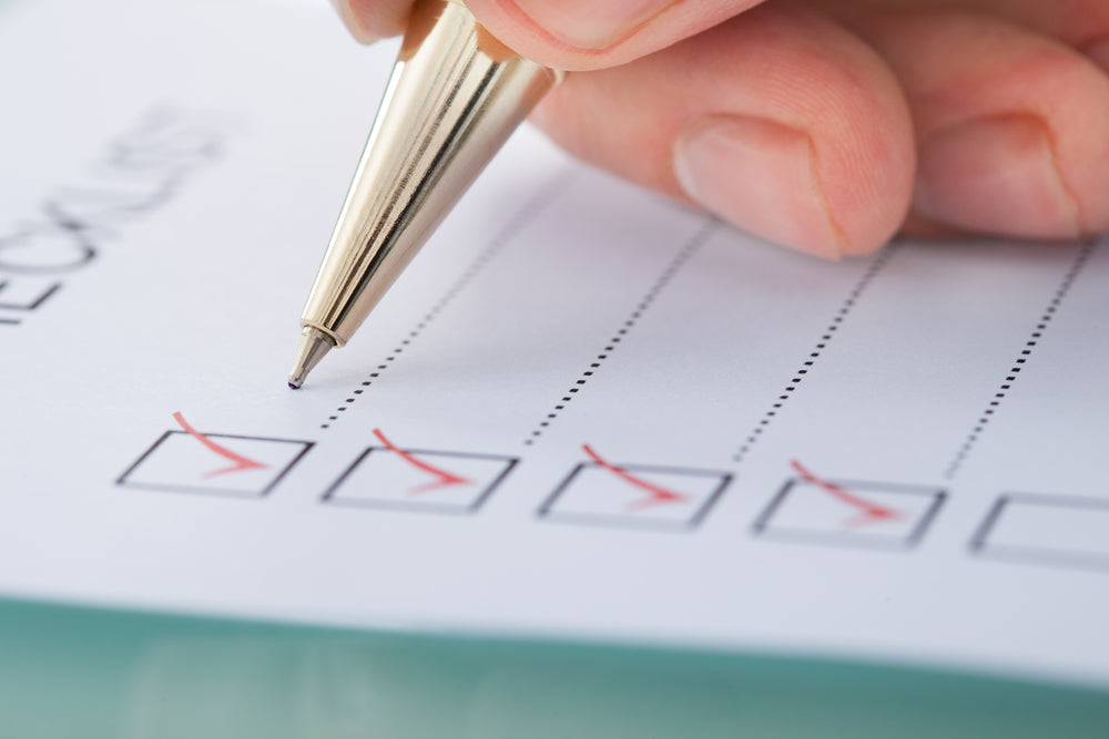 A hand holds a gold pen, marking off items on a checklist printed on white paper, indicating preparation tasks related to post weight-loss surgery.