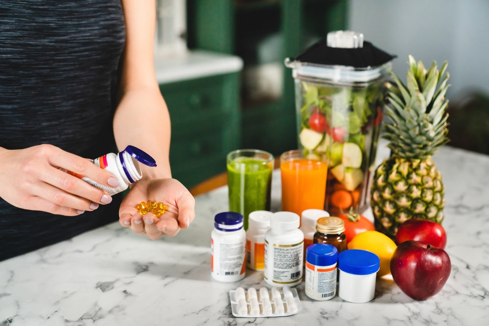 A person in a black tank top pours yellow capsules from a bottle into their palm, surrounded by colorful fruits, juices, and various nutrition supplements on a marble countertop.