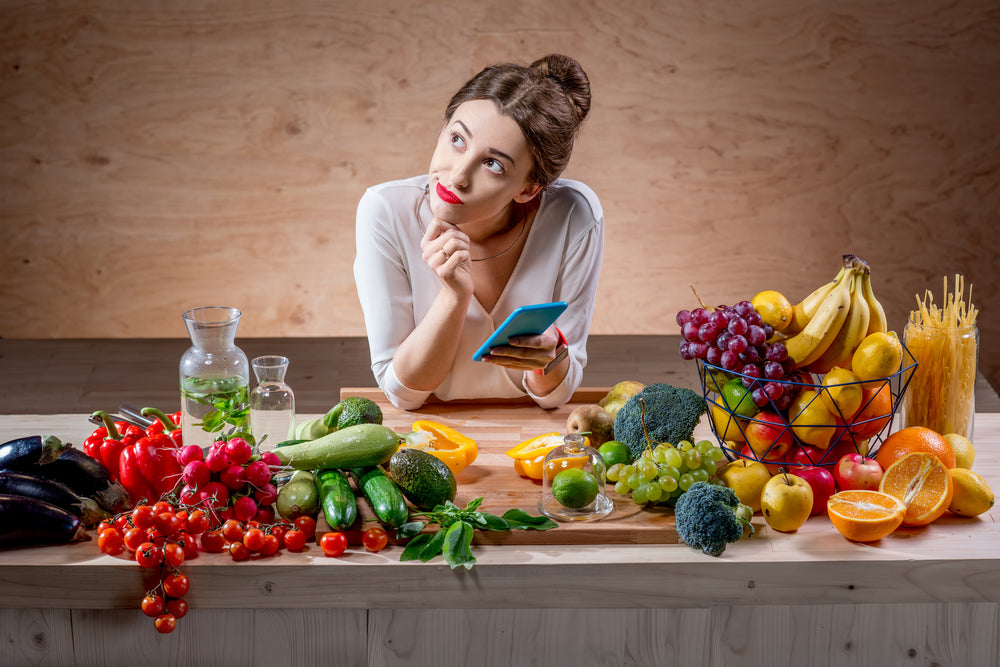 A woman with a thoughtful expression holds a blue smartphone while surrounded by a variety of colorful fruits and vegetables on a wooden table, suggesting a focus on healthy eating and nutrition.