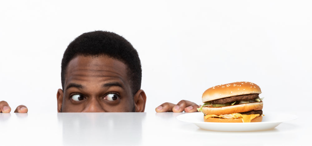 A man peeks over a table with a curious expression, looking at a large burger on a plate in front of him, illustrating the struggle with hunger and cravings after surgery.
