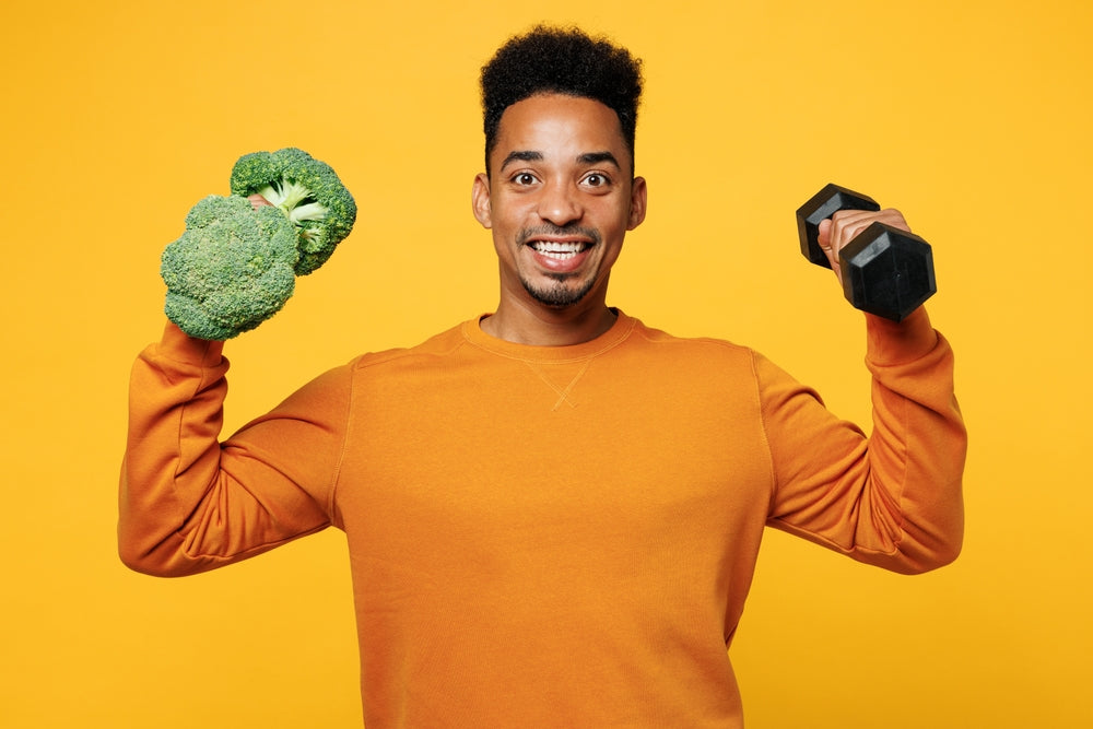 A smiling man in an orange sweatshirt holds a piece of broccoli in one hand and a dumbbell in the other against a bright yellow background, promoting healthy living and fitness.