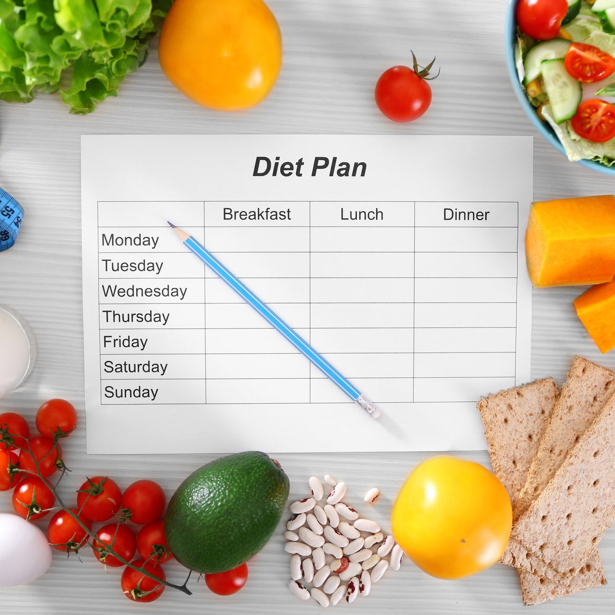A blank diet plan sheet with days of the week is surrounded by fresh vegetables, fruits, and grains, highlighting healthy food options for a bariatric surgery diet.