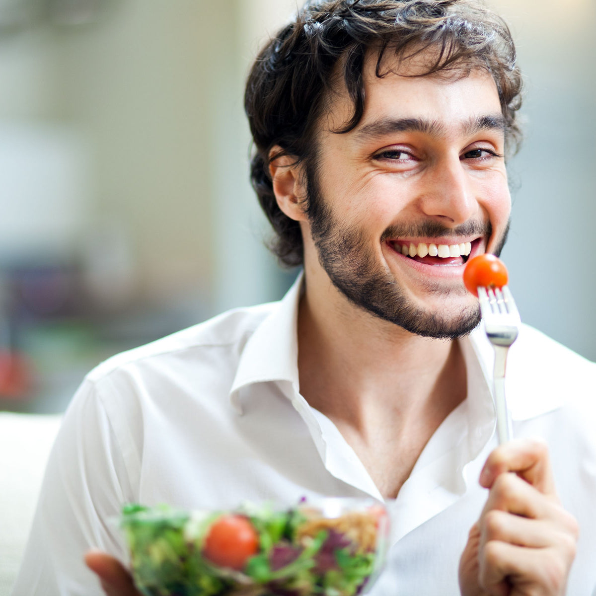 A smiling man in a white shirt holds a fork with a cherry tomato, ready to eat, while sitting with a bowl of salad in a bright, casual indoor setting.