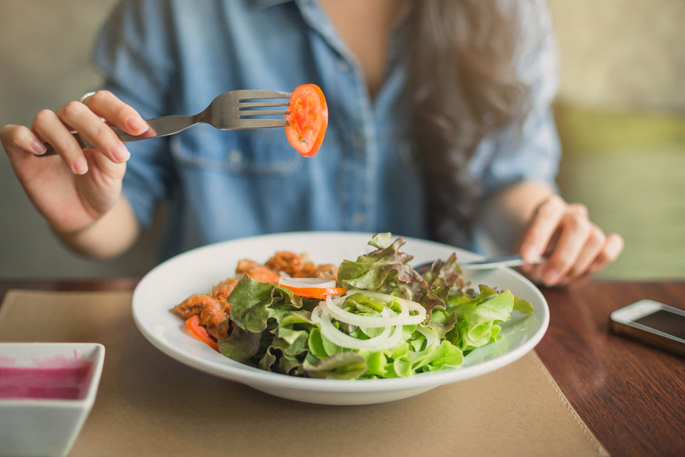 A person in a denim shirt holds a fork with a slice of tomato above a fresh salad in a bowl, sitting at a wooden table with a smartphone nearby, emphasizing healthy eating choices.