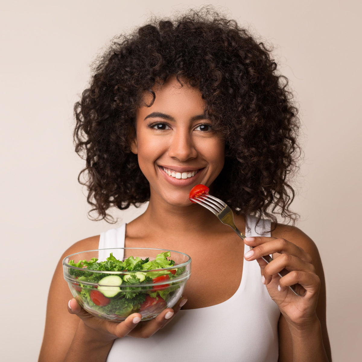 A smiling woman with curly hair holds a bowl of salad in one hand and a fork with a cherry tomato near her mouth, set against a neutral background, promoting healthy eating for weight loss.