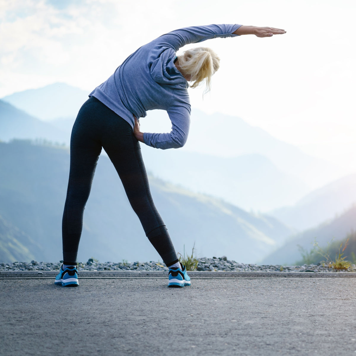 A woman in athletic wear stretches to the side against a scenic mountain backdrop, promoting movement and exercise after bariatric surgery.