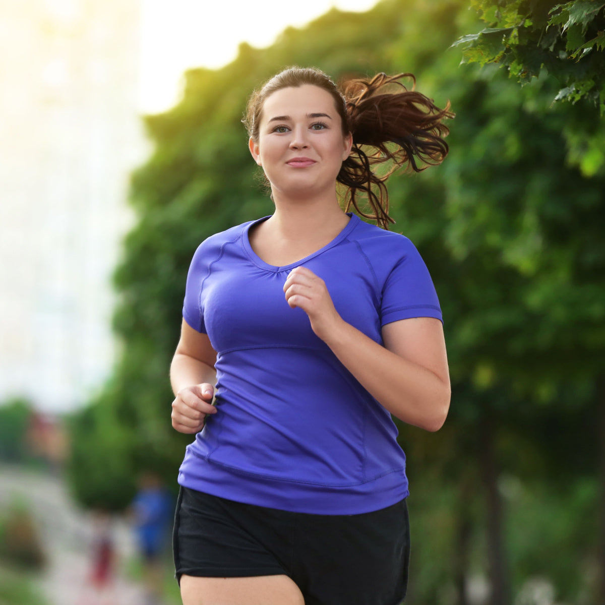 A young woman with a joyful expression runs along a tree-lined path, wearing a purple athletic shirt and black shorts, embodying an active lifestyle after bariatric surgery.