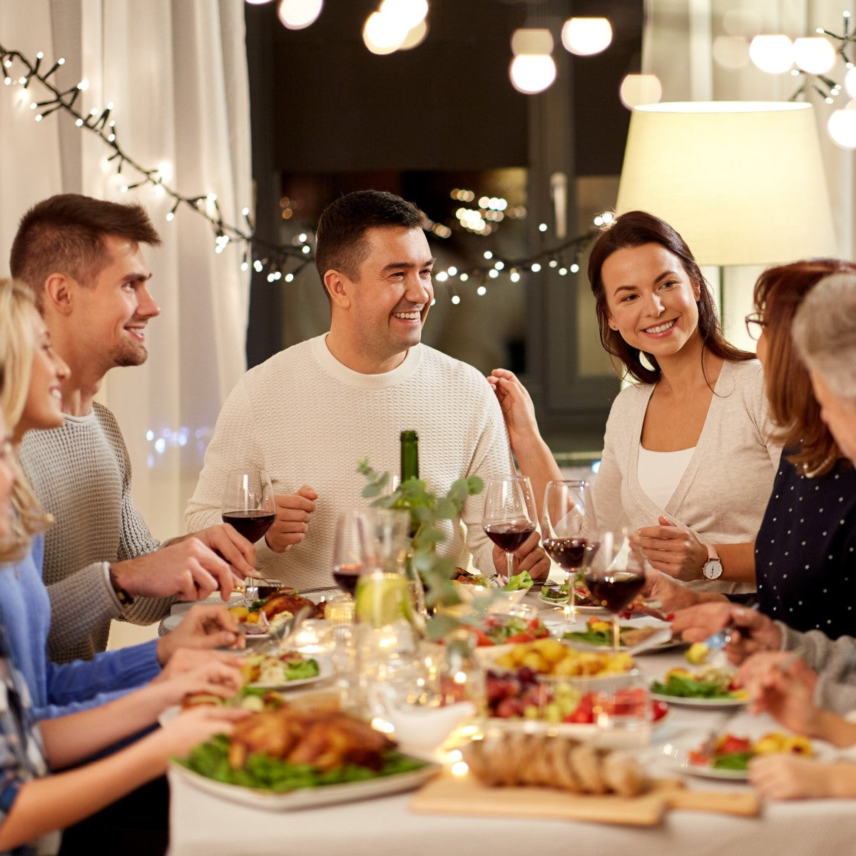 A group of six people enjoy a festive dinner at a table filled with food and wine, smiling and engaging in conversation under warm, ambient lighting with holiday decorations.
