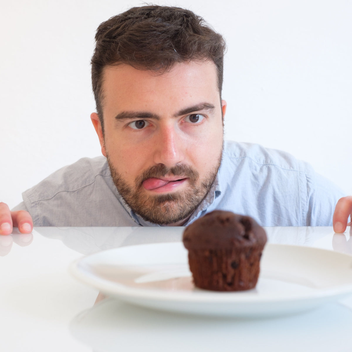 A man with a beard and short hair leans forward with a focused expression and his tongue out, staring at a chocolate muffin on a white plate in a bright, minimalistic setting.