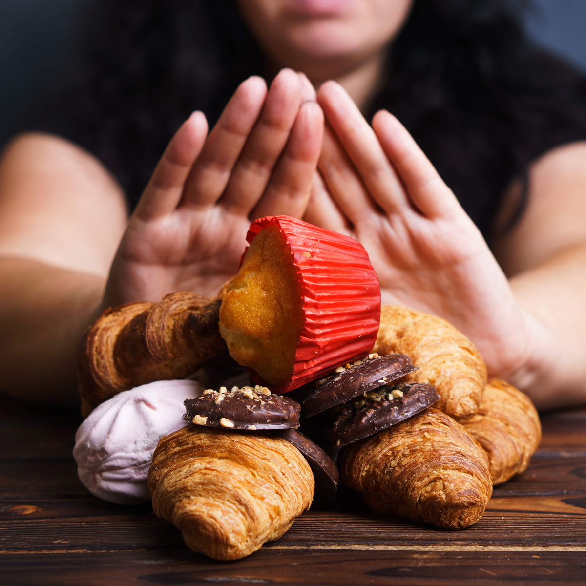 A person with curly hair holds their hands up in a stopping gesture in front of a pile of pastries, including croissants, cookies, and a muffin, symbolizing foods to avoid after bariatric surgery.