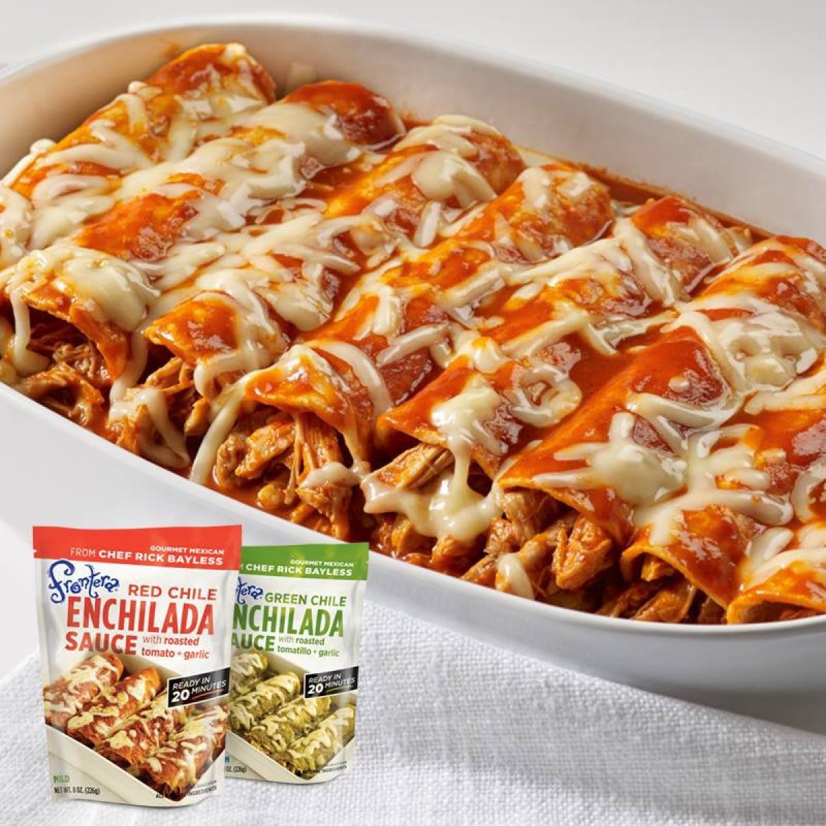 A white dish holds several enchiladas topped with melted cheese and red sauce, accompanied by two packets of Frontera enchilada sauces, showcasing a delicious Mexican meal.