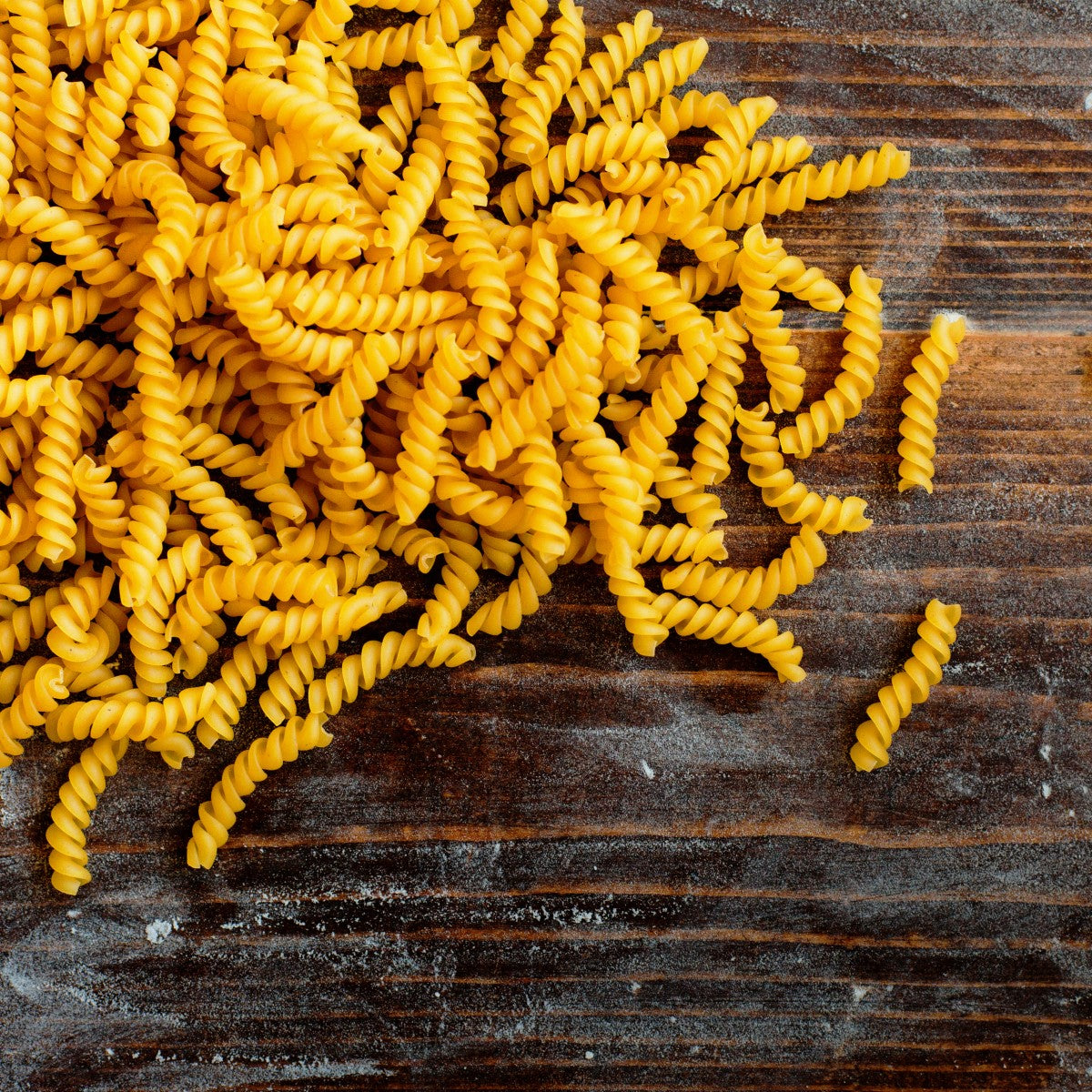 A pile of yellow fusilli pasta is scattered on a wooden surface, showcasing its twisted shape and texture against the rustic backdrop.