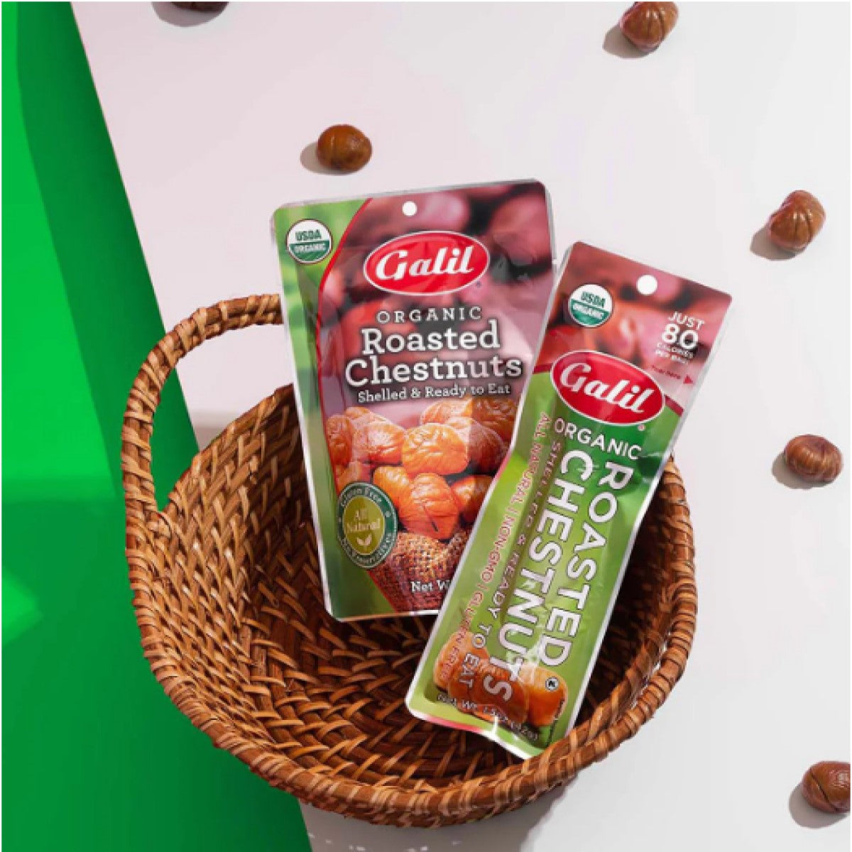 Two packages of Galil organic roasted chestnuts, one in a green and red design and the other in a clear pouch, are placed in a woven basket on a bright background with scattered chestnuts.