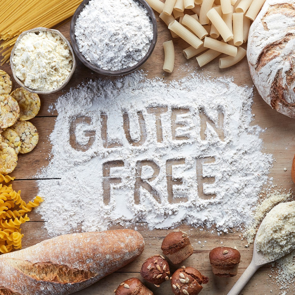 A wooden surface is covered with various gluten-free products, including pasta, flour, and baked goods, with the words "GLUTEN FREE" written in flour.