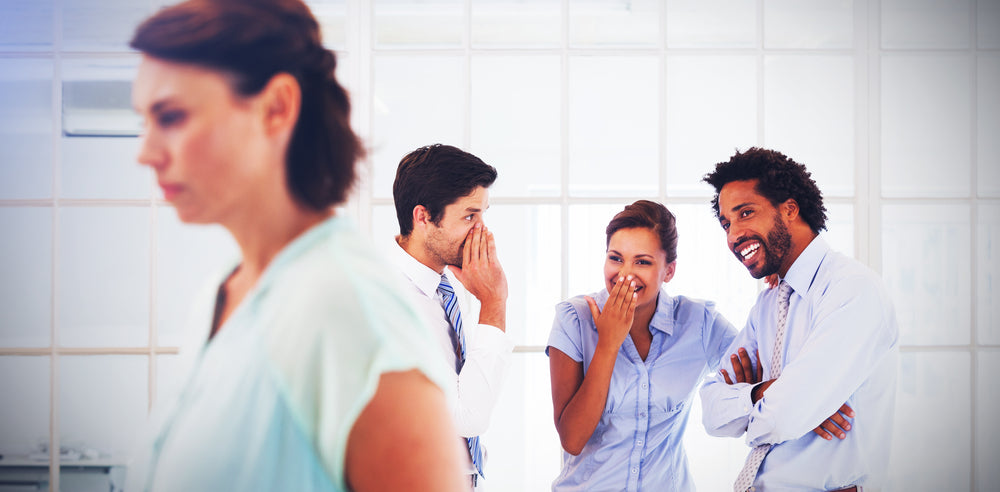 A woman looks upset while walking away from a group of three colleagues, who are whispering and laughing together, in a bright office setting with large windows.