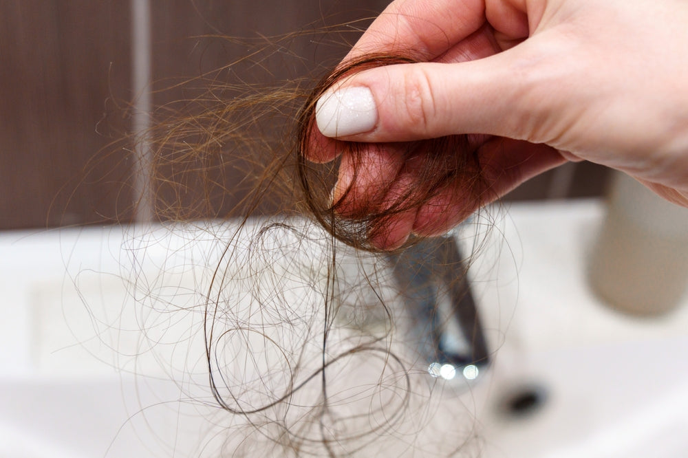 A hand holds a clump of brown hair above a bathroom sink, with a blurred faucet and countertop in the background, illustrating concerns about hair loss after bariatric surgery.
