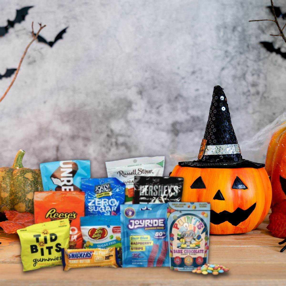 A festive Halloween display features a smiling orange pumpkin wearing a black hat, surrounded by various snacks including candies and treats, set against a backdrop of bats and autumn decorations.