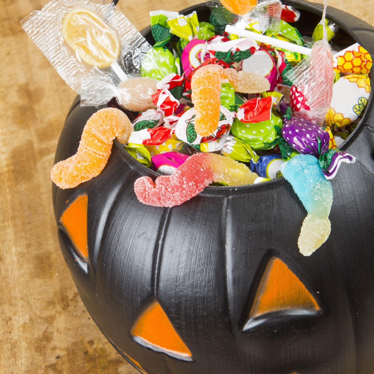 A black pumpkin-shaped bowl filled with colorful Halloween candies, including gummy worms and lollipops, sits on a wooden surface, showcasing bariatric-friendly treats for the holiday.