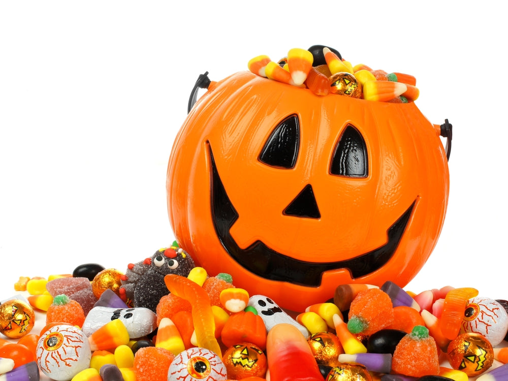 A smiling orange jack-o'-lantern is filled with colorful candy and surrounded by various Halloween treats, creating a festive and playful Halloween scene.