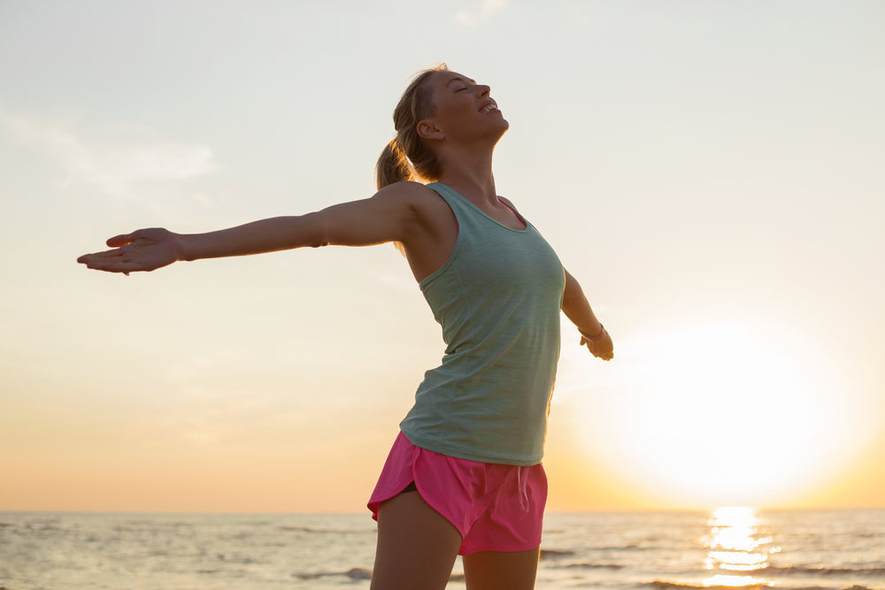 A woman stands on a beach at sunset, arms outstretched and head tilted back, expressing joy and freedom after achieving weight loss success.