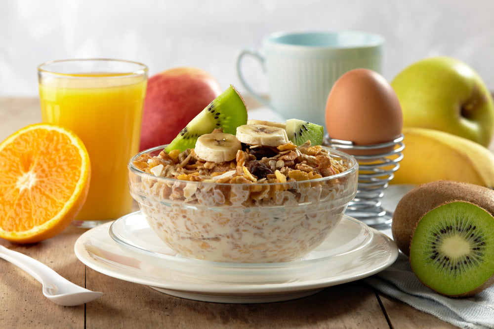 A bowl of oatmeal topped with banana slices and kiwi sits on a plate, surrounded by a glass of orange juice, an egg in an egg cup, and various fruits on a wooden table.