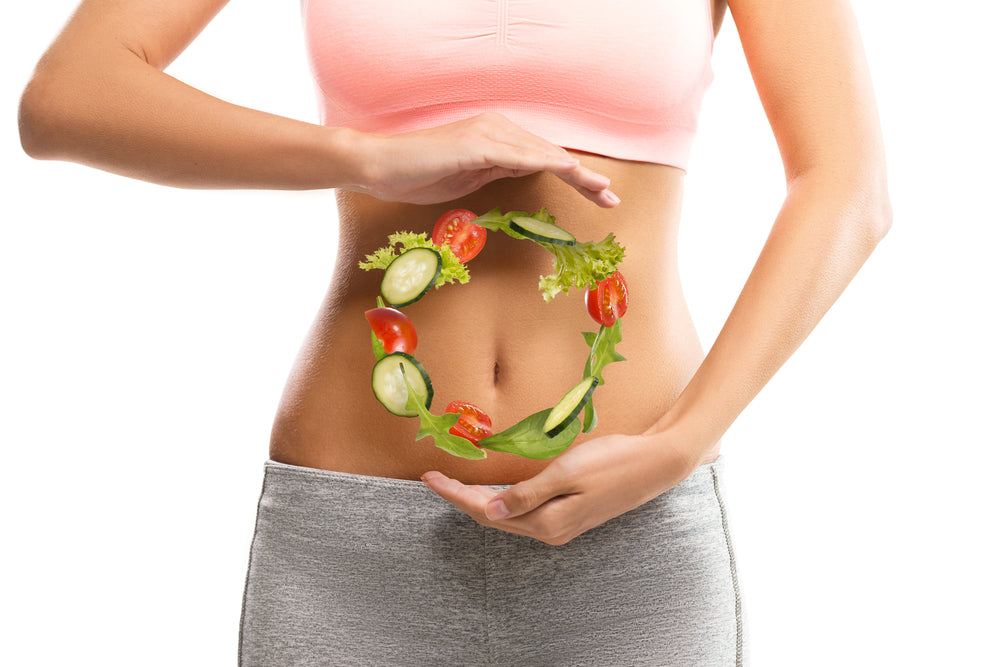 A person in a pink sports bra holds their hands around a circular arrangement of fresh vegetables, symbolizing healthy eating and wellness, against a bright, plain background.