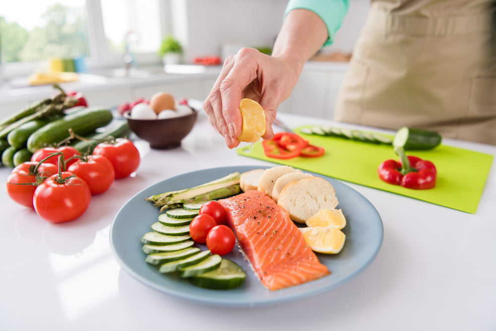 A person squeezes lemon over a plate of salmon, surrounded by fresh vegetables like tomatoes, cucumbers, and asparagus, in a bright kitchen setting.