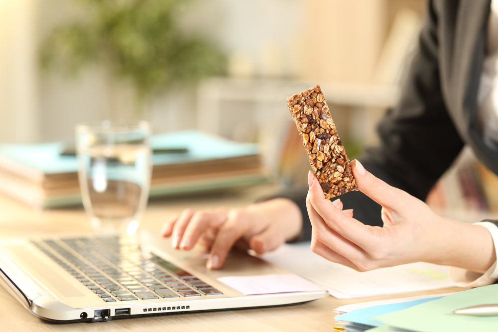 A person in business attire holds a granola bar in one hand while typing on a laptop with the other, surrounded by office supplies and a glass of water on a desk.
