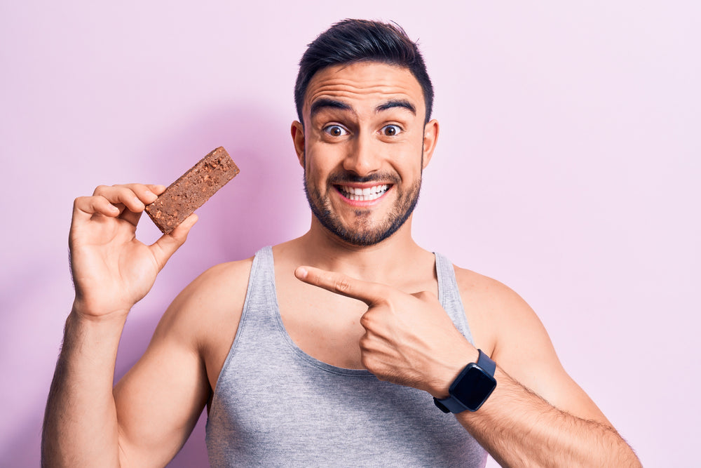 A smiling man in a gray tank top holds a protein bar in one hand and points to it with the other, against a light pink background, promoting healthy snacking options.
