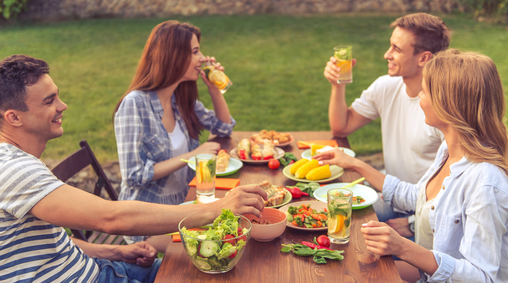 Four friends enjoy a lively outdoor meal at a wooden table, sharing healthy dishes like salads and grilled vegetables while laughing and toasting with drinks in a sunny garden setting.